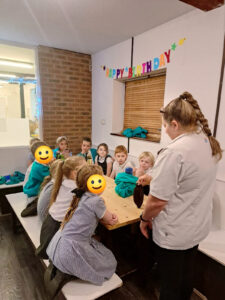 Some Year 4 pupils are photographed sat around a long table together, listening to a staff member from Temper Temper Chocolate who is speaking to them.