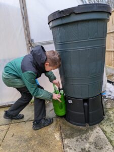 A young boy is seen wearing his winter coat and bending down to fill up his watering can from a water butt in a large greenhouse.