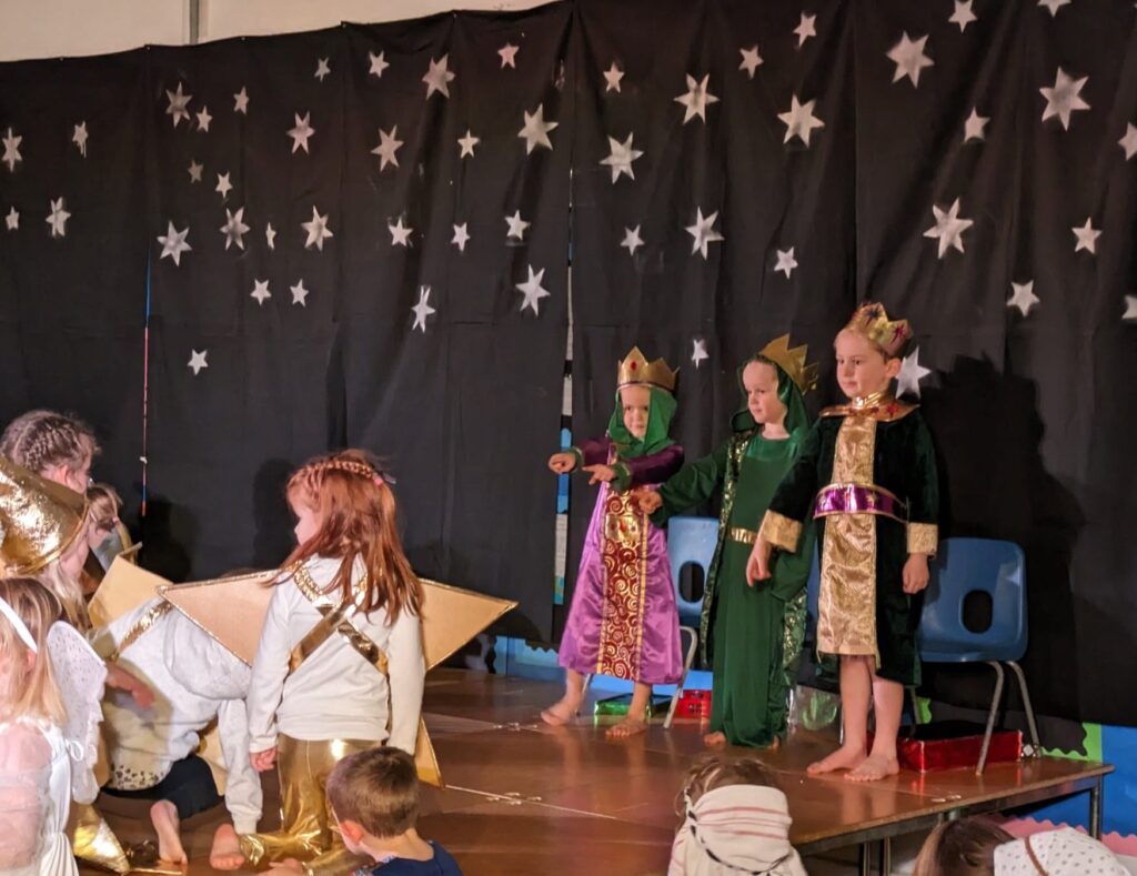 A photo of three young girls standing on stage in the main hall in Nativity clothing during a school performance.