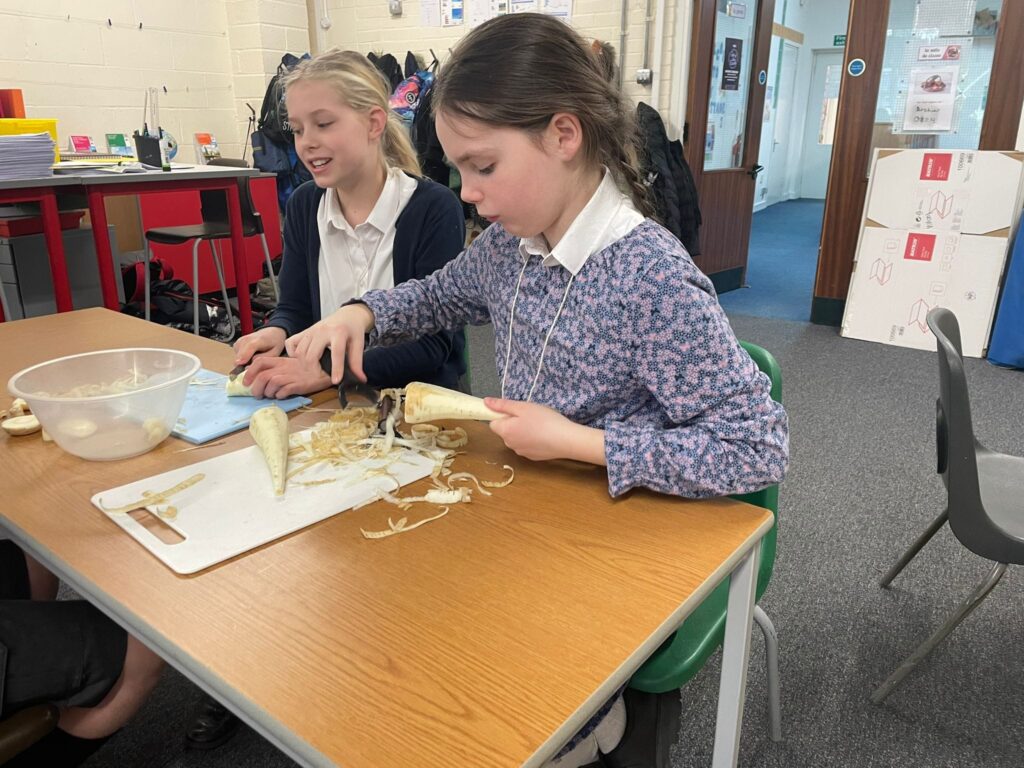 Two female students are pictured sat together at a desk, dressed as evacuees, peeling vegetables.