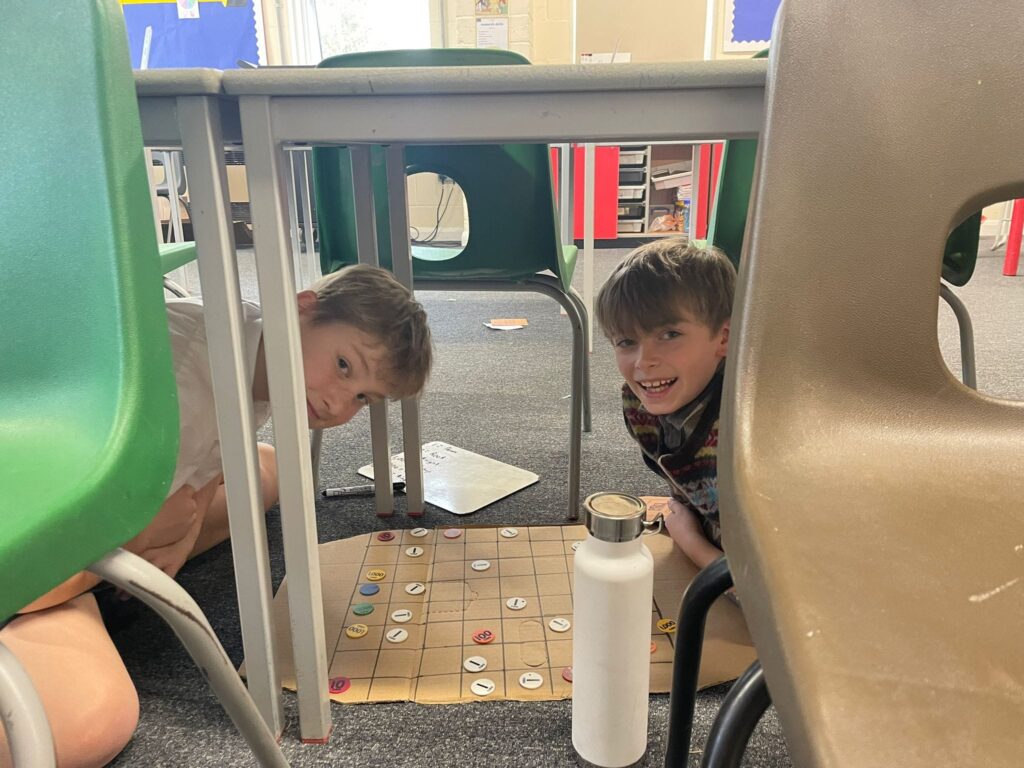Two male students are pictured sitting under a desk in their classroom, wearing WWII evacuee-style clothing. They are seen playing a board game and smiling for the camera.