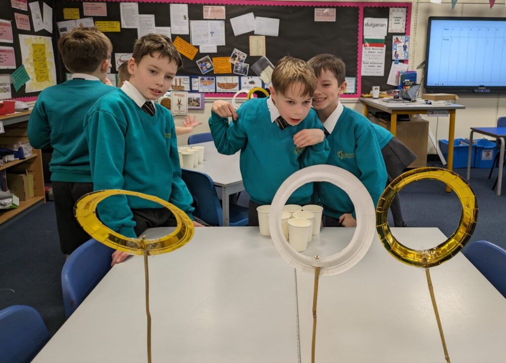 Three male students are pictured smiling together, as they practice throwing balls through large hoops on sticks they have created.