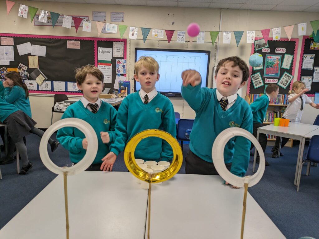 Three male students are seen throwing balls through small hoops on sticks on the table in front of them.