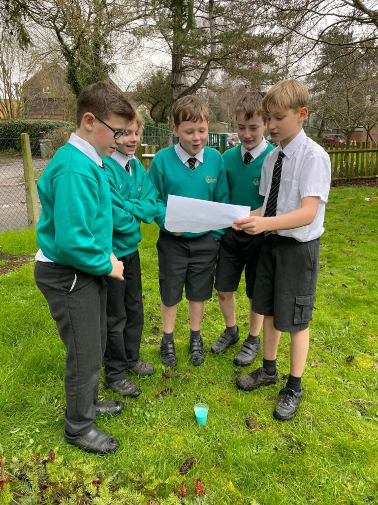 A group of five male students are pictured outdoors on a grassy area, reading from a sheet of paper and a 'potion' they have created together.