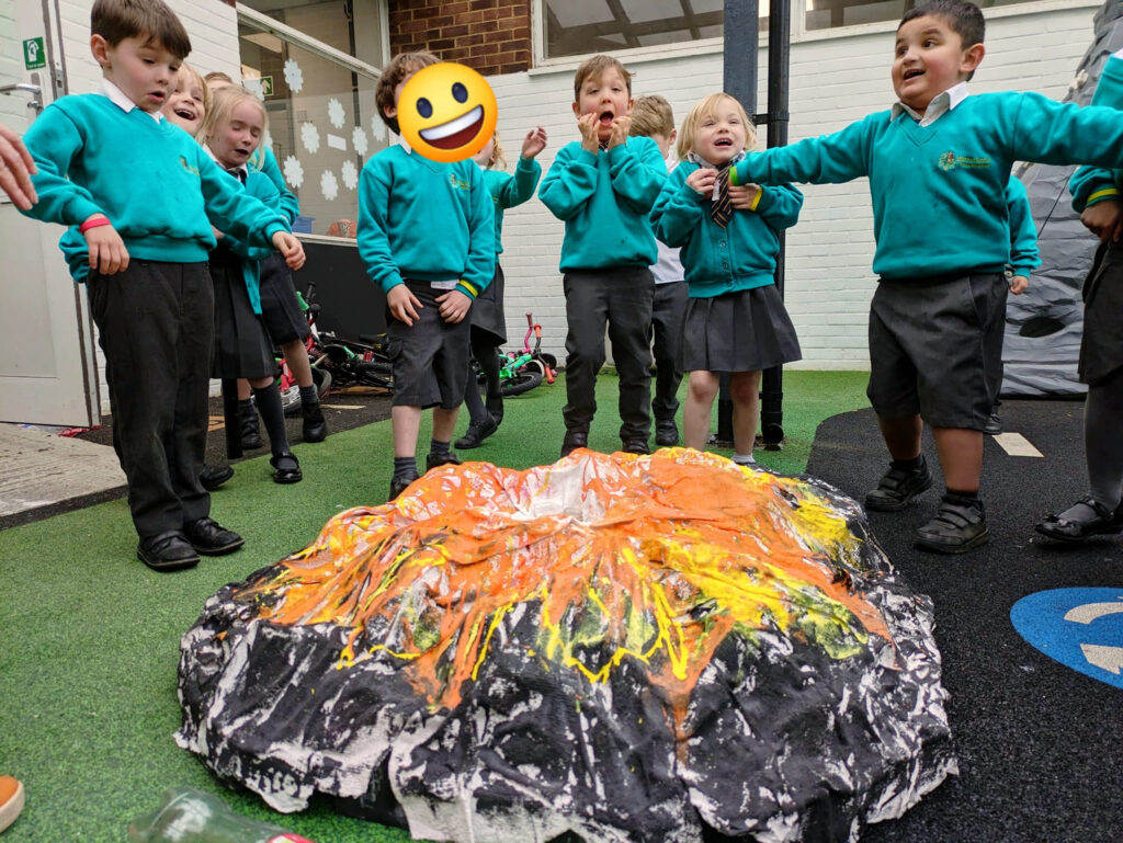 A small group of EYFS pupils are pictured smiling together, whilst looking at a model of an erupting volcano they have built.