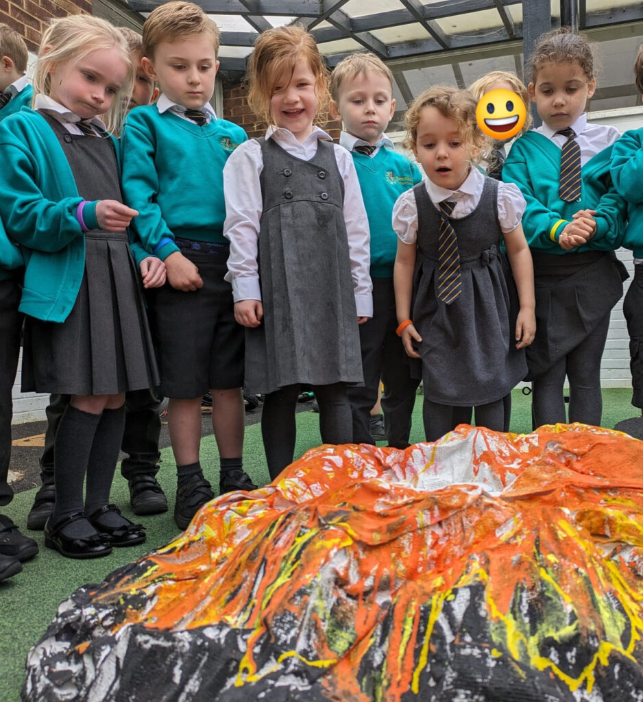 A small group of EYFS pupils are pictured smiling together, whilst looking at a model of an erupting volcano they have built.