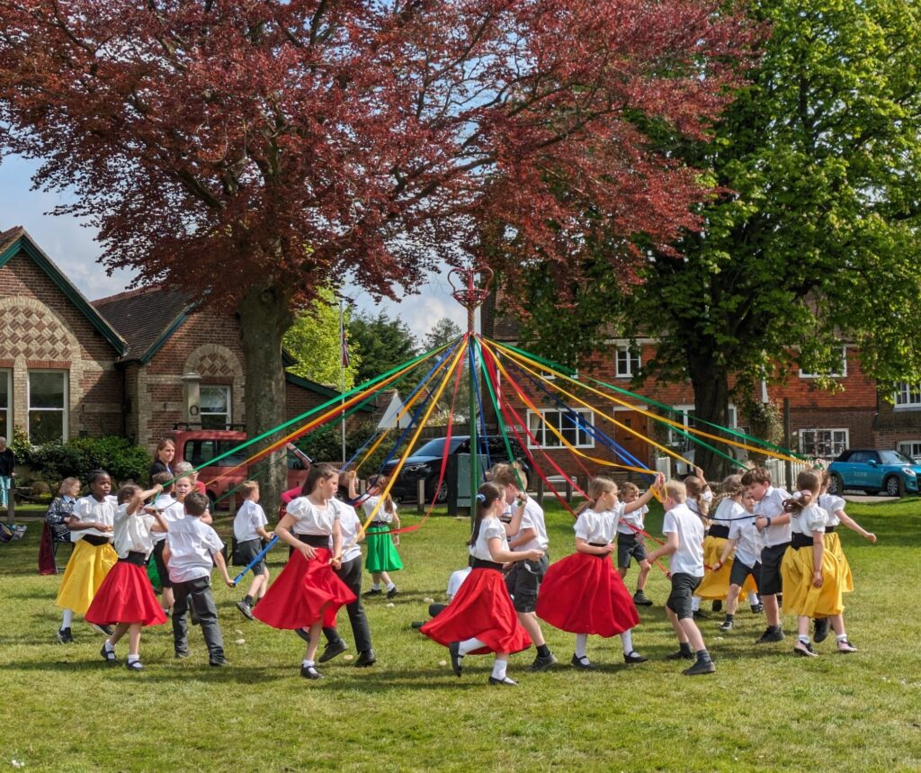 Photo showing a group of Horsmonden pupils dressed in bright-coloured clothes, dancing together around a maypole to celebrate May Day 2024.
