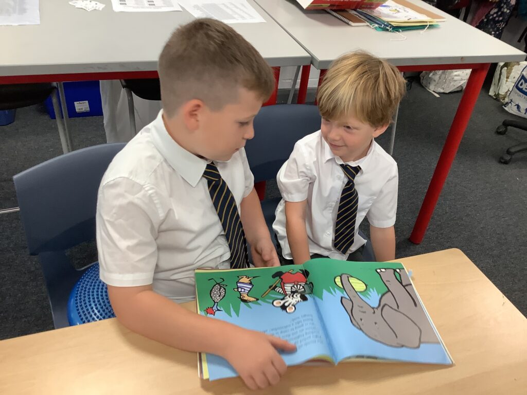Two boys in school uniform sitting near a desk, looking at each other over an open picture book.