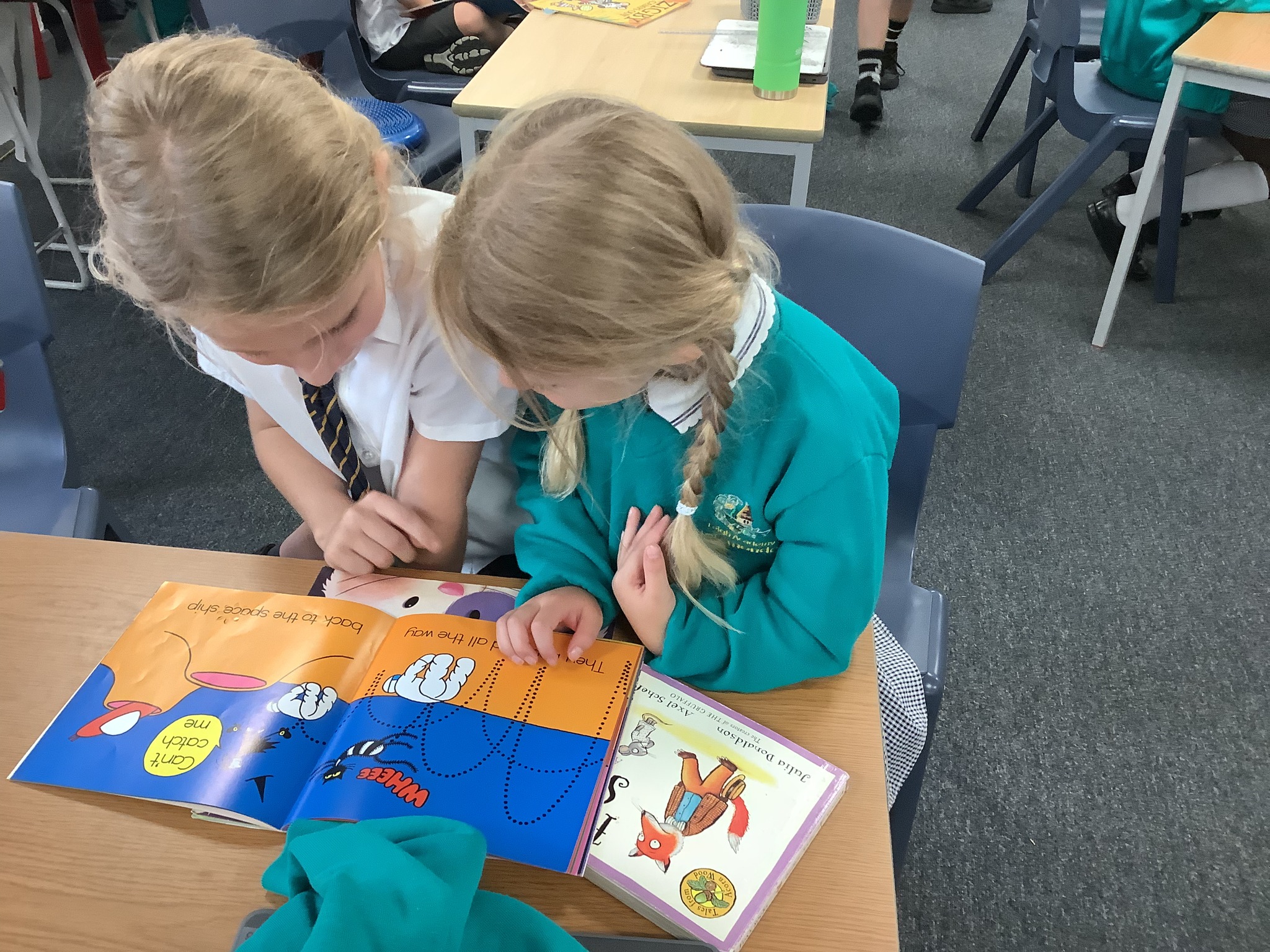 Two girls in school uniform sitting at a desk and reading an open picture book together.