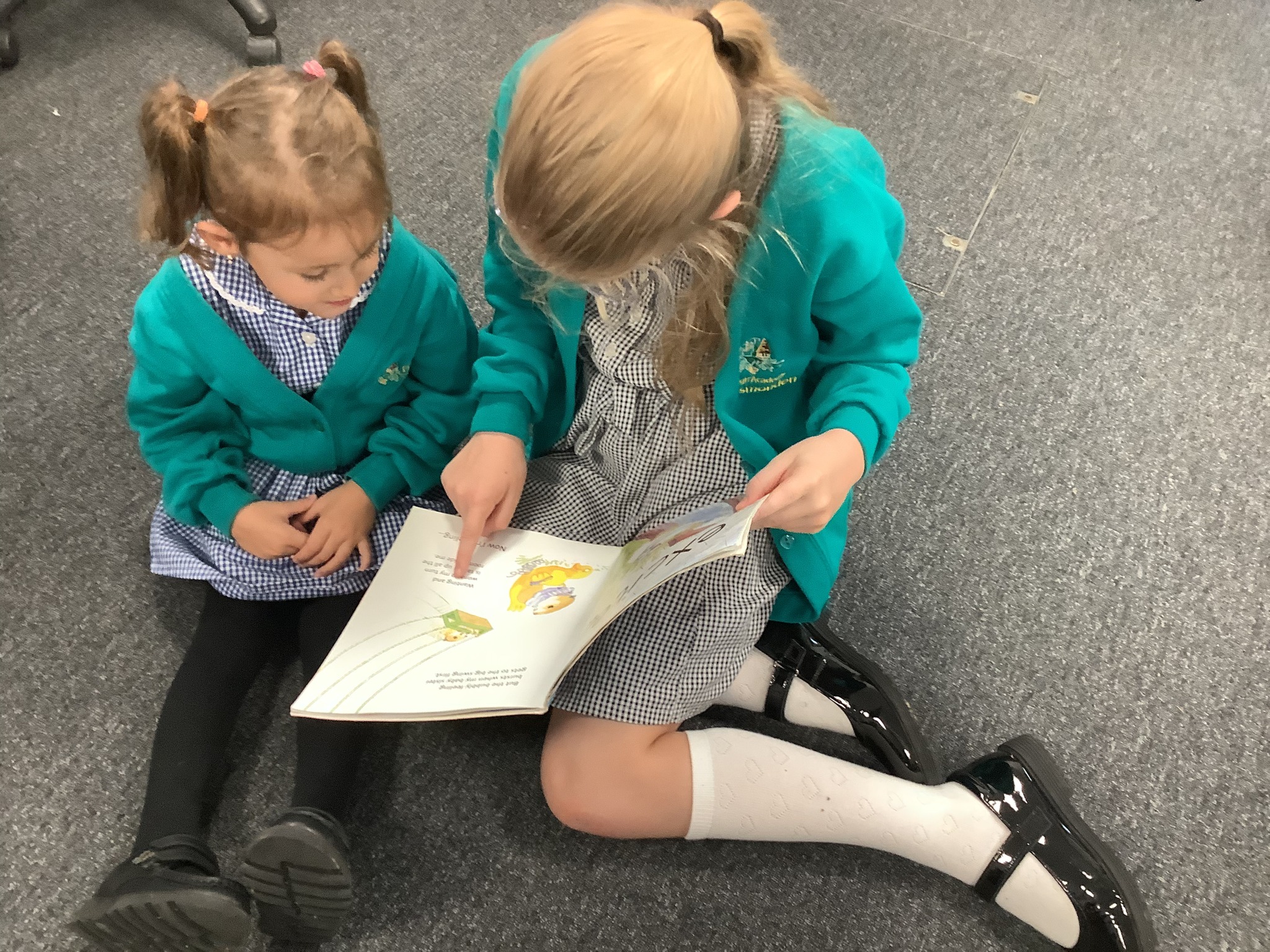 Two girls in school uniform sitting on a grey carpet, looking at an open picture book.