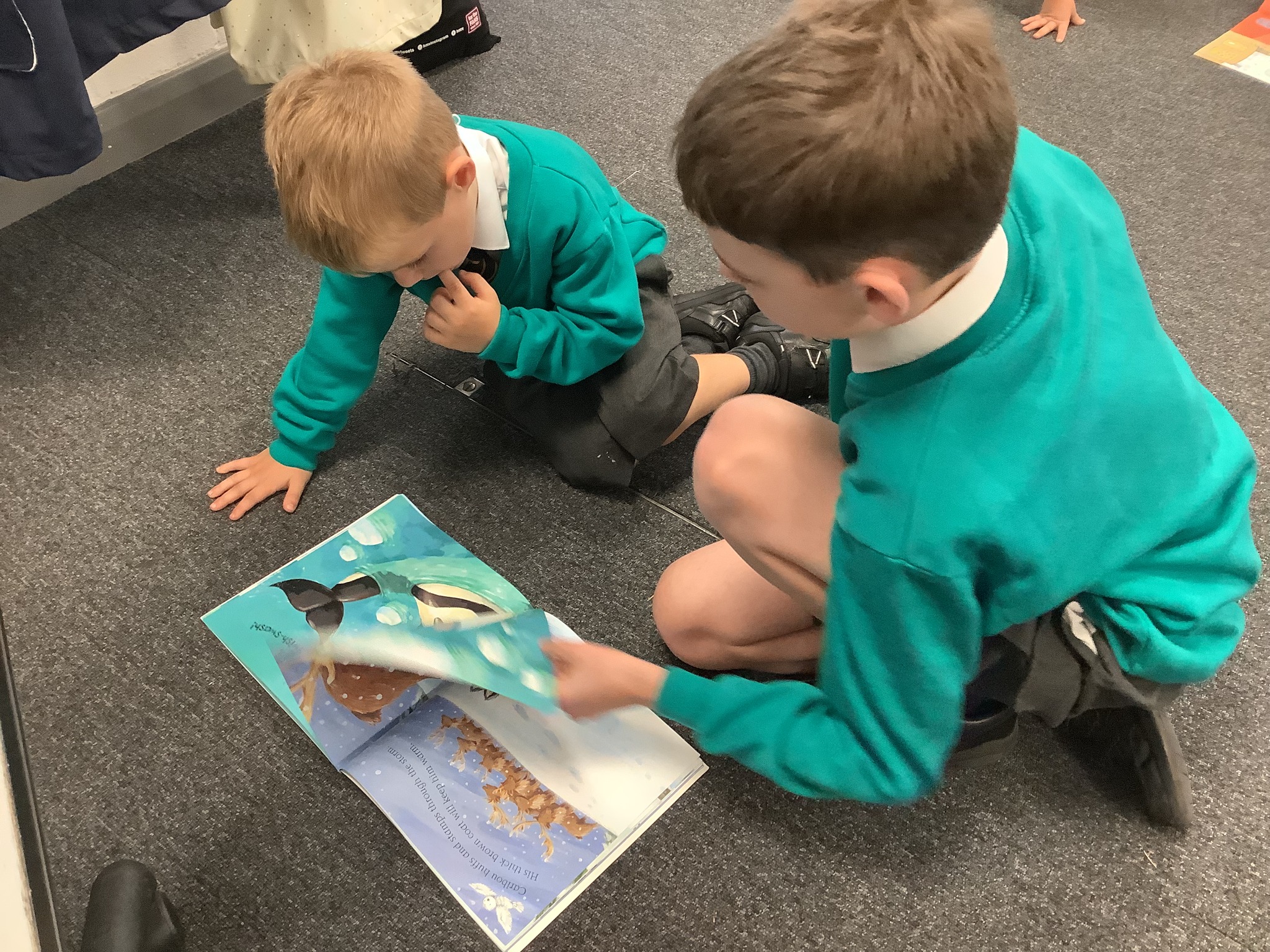 Two boys in school uniform kneeling on the floor, looking at an open picture book.