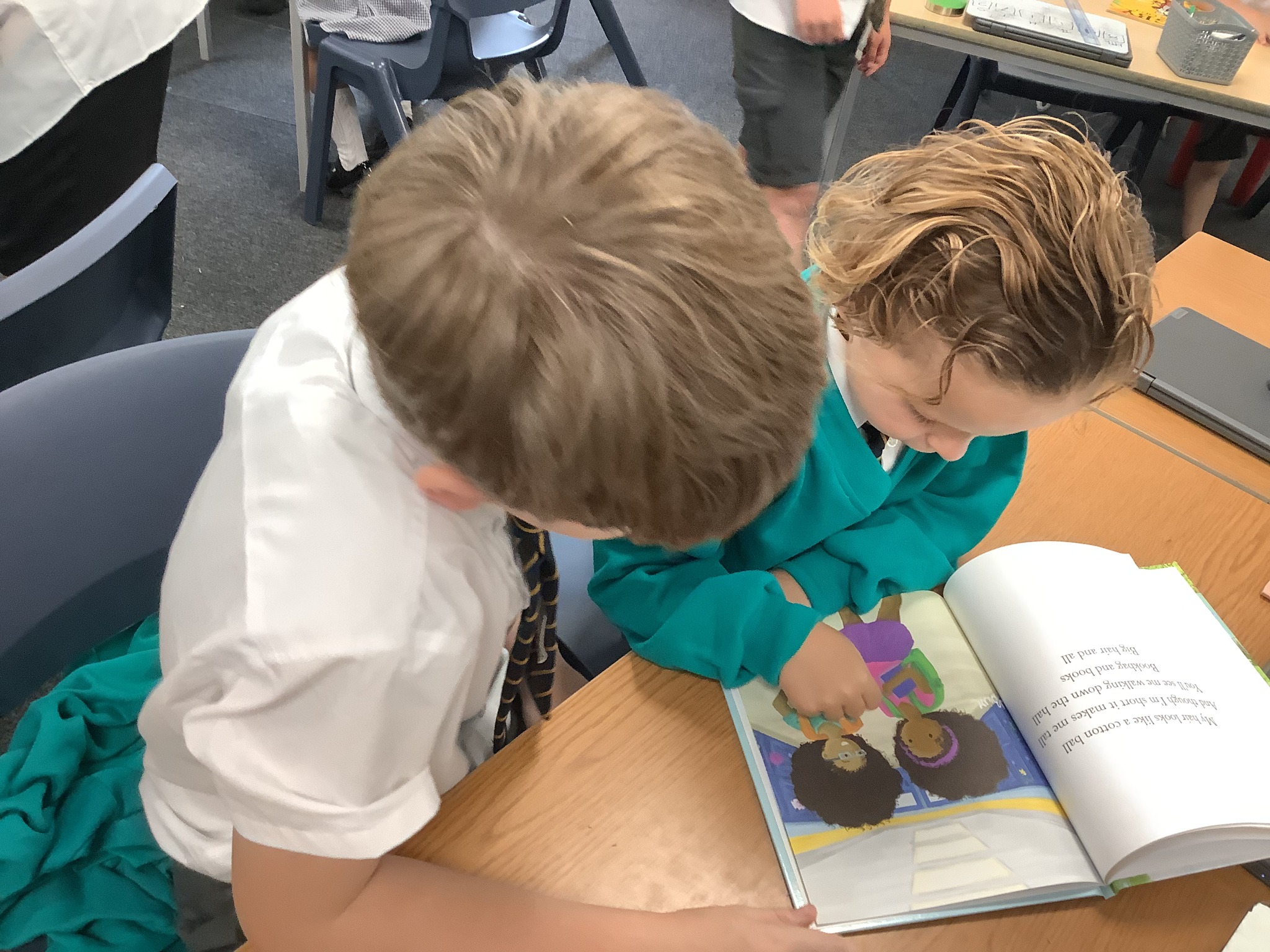 Two children in school uniform looking intently at an open picture book on a wooden desk.
