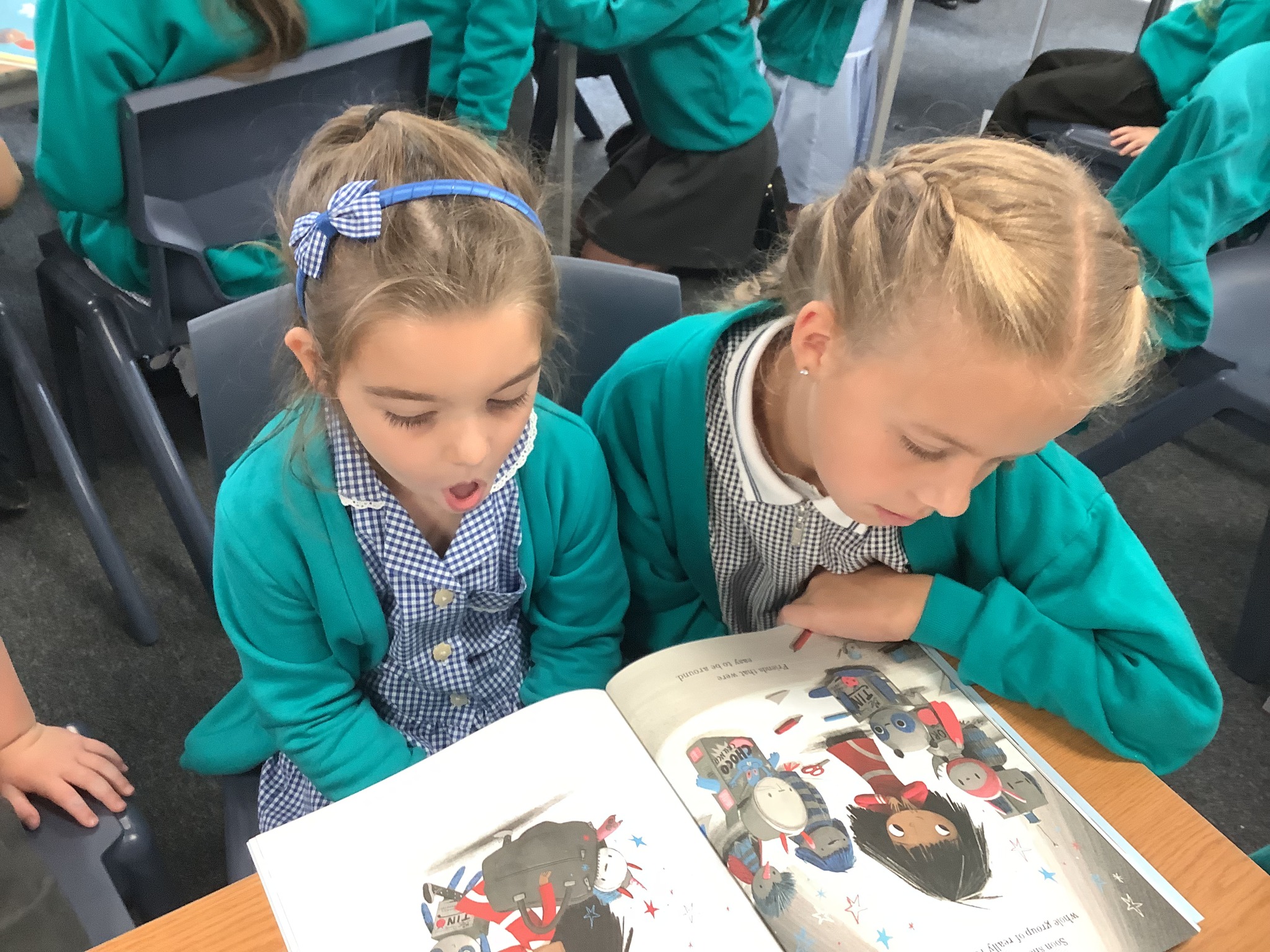 Two girls in school uniform, sitting at a desk and engrossed in reading an open picture book.