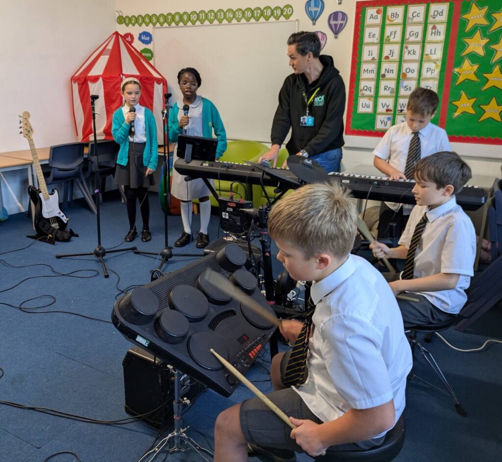 A teacher and pupils in band practice. Two are playing electric drums and one is using a keyboard with two singing.