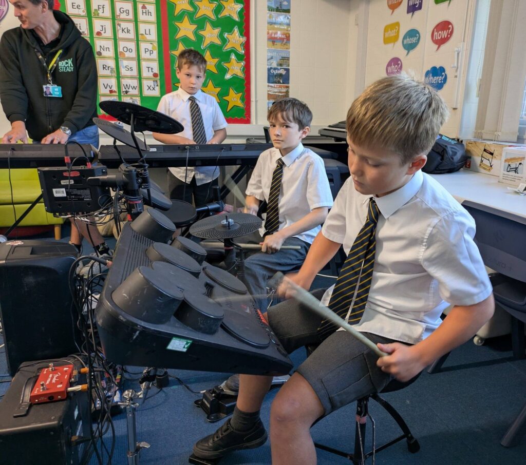 Three pupils in a band practice. Two are playing electric drums and one is using a keyboard