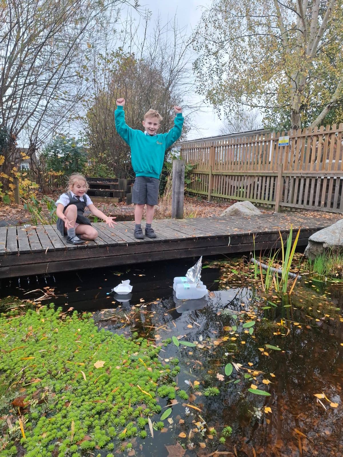 Two children on a wooden deck next to a pond, one kneeling and one standing with hands raised in excitement, watching their boats float.