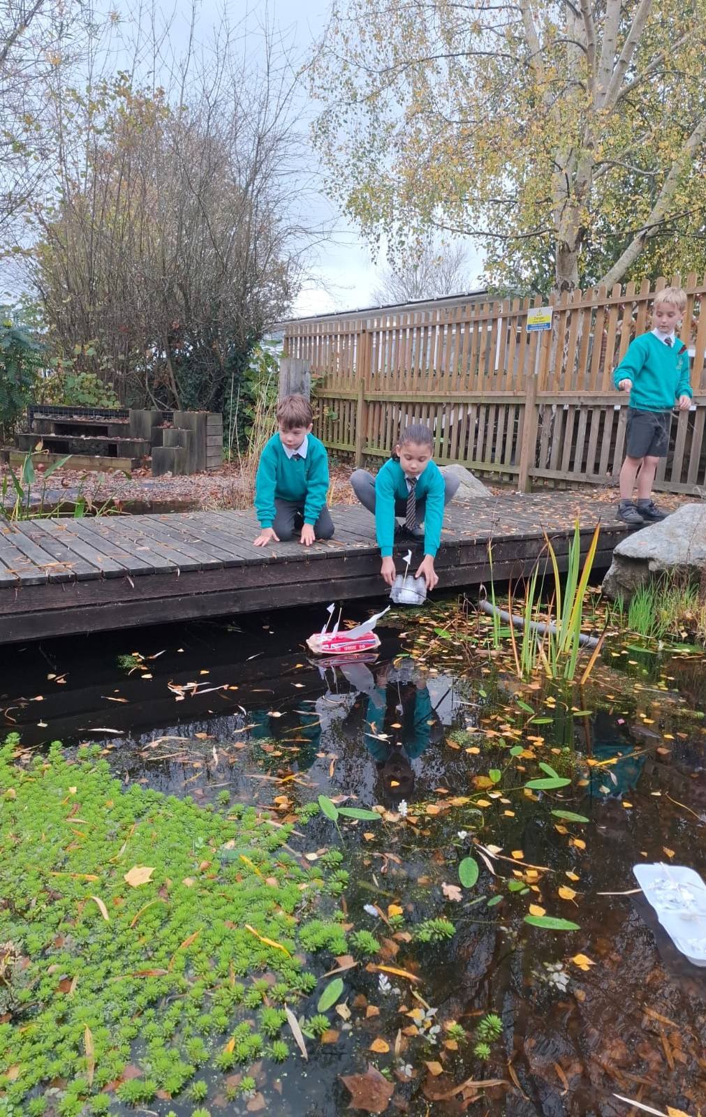 Two young boys kneeling on a wooden deck next to a pond, launching a red and white boat into the water.
