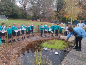 Group of children in school uniforms stand around an outdoor pond, watching a teacher launch their homemade boats.