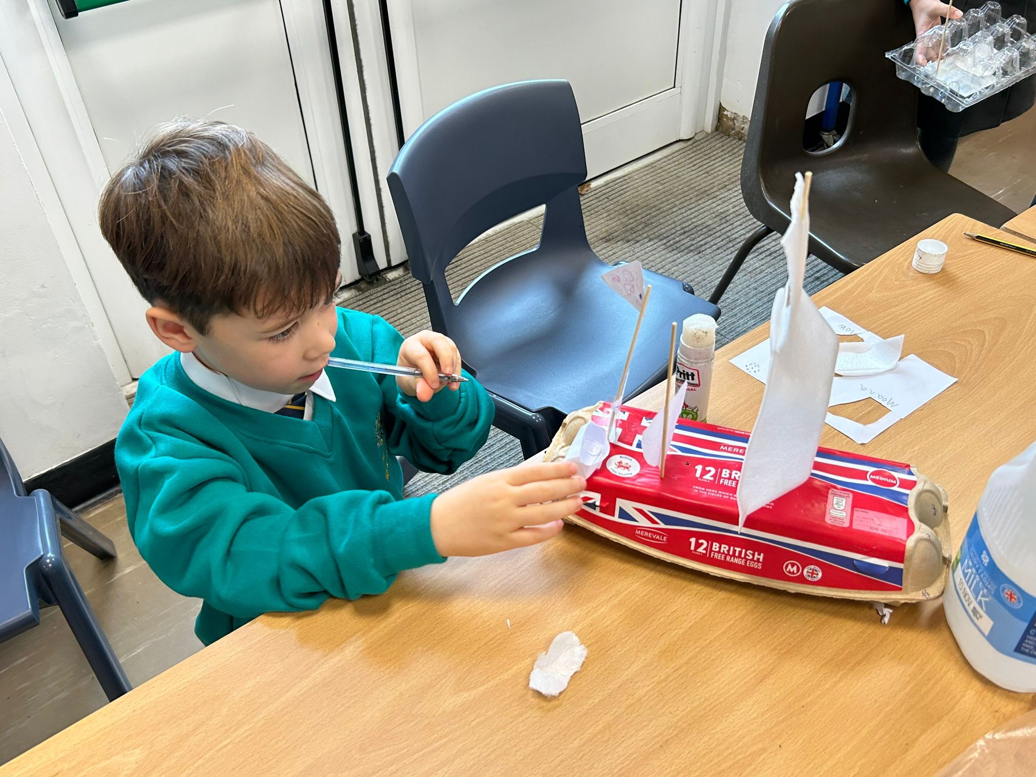 Boy in a green jumper working on a model boat made from a red and white cardboard box, adding a paper sail with a pen.