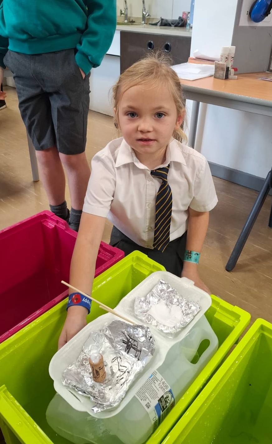 Young girl in school uniform stands over water tubs, holding a homemade foil and polystyrene boat containing a small figure and white powder.