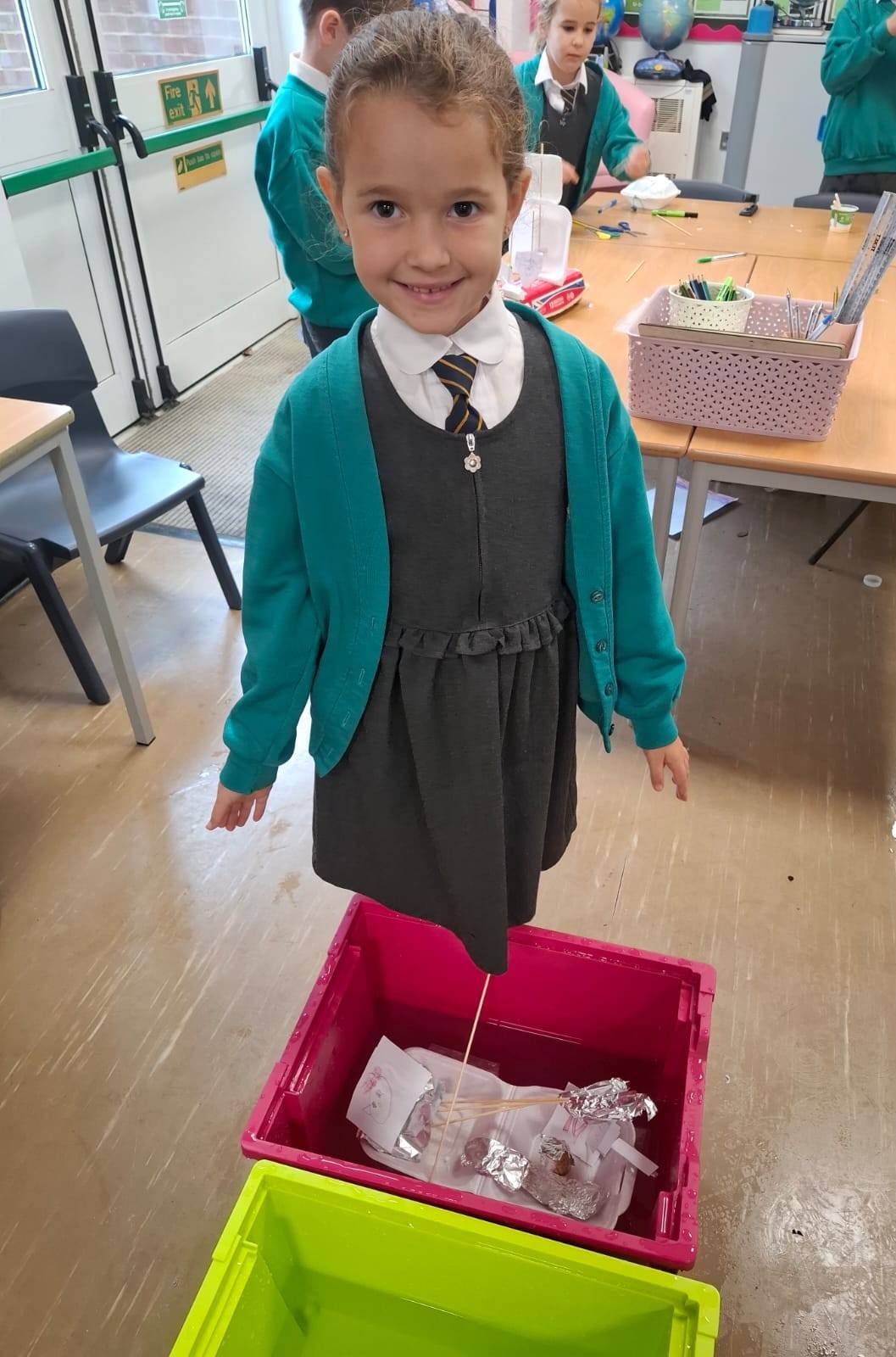 Young girl in a school uniform and turquoise cardigan smiling, standing over a pink tub of water containing her homemade foil boat.
