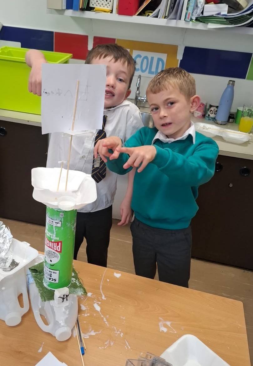 Two young boys in a classroom holding up a boat made from a Pringles can base and a paper sail on sticks.