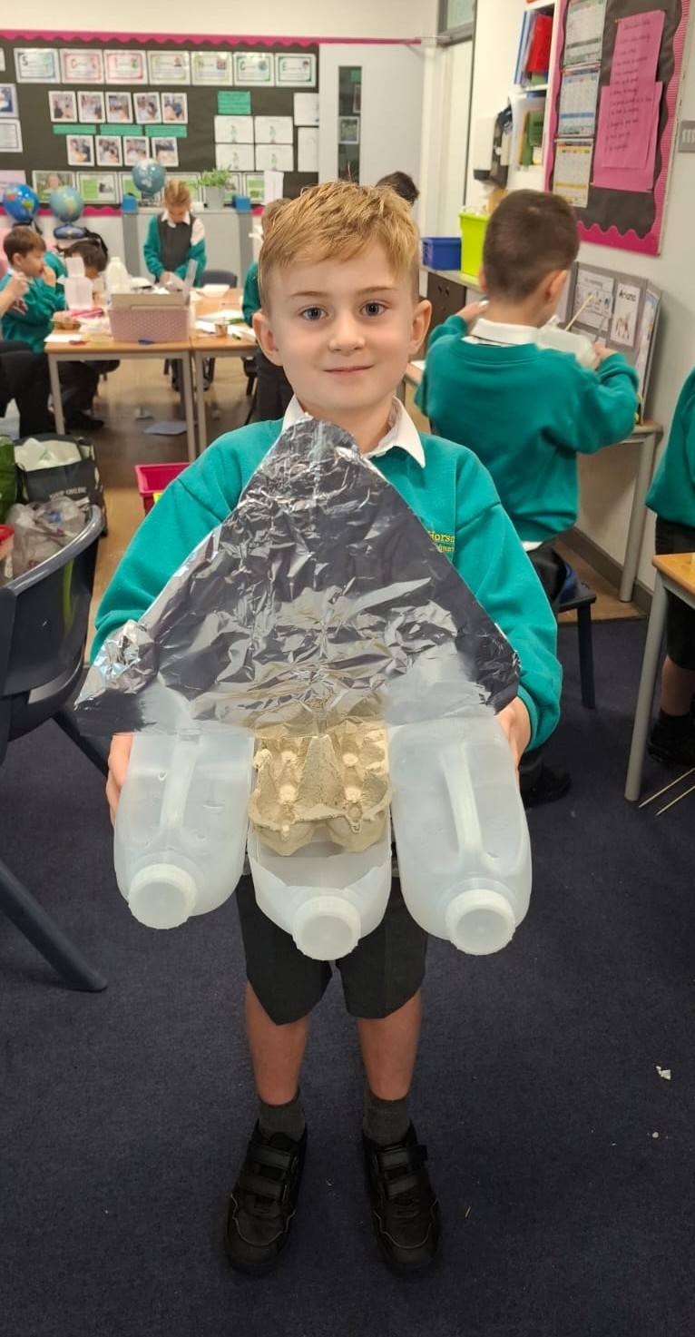 Boy in a green jumper holding up a large homemade boat made from three plastic milk jugs, an egg carton, and a foil cover.