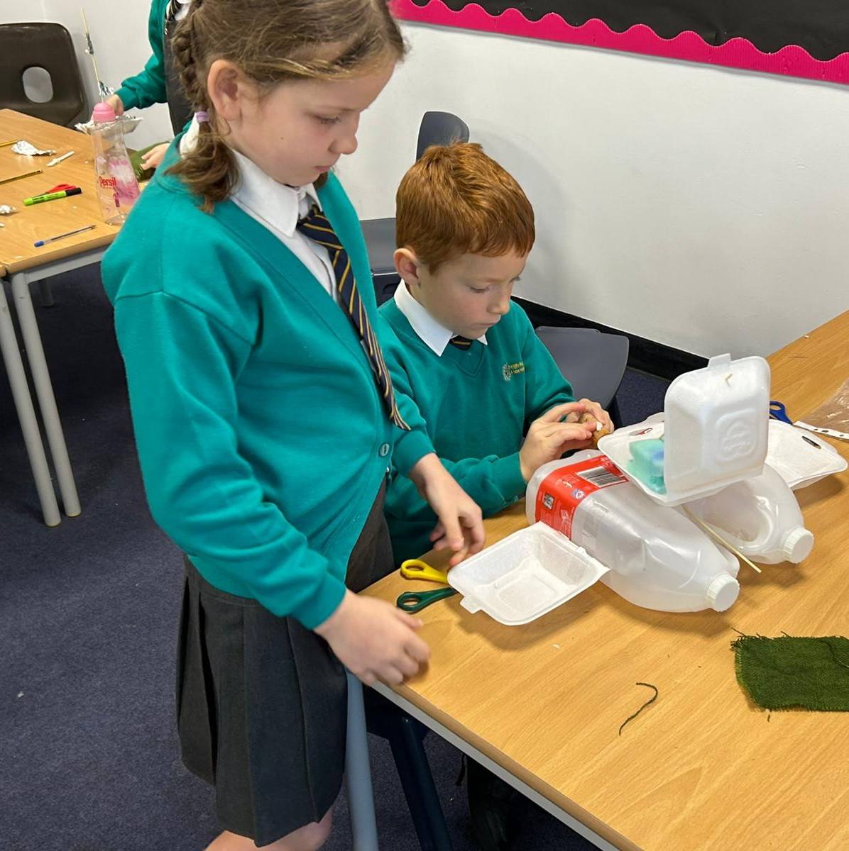 Boy and girl in green jumpers working on a model boat made from a red and white cardboard box, adding a boat made out of plastic bottles and polystyrene packaging