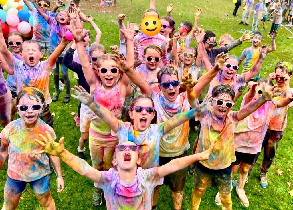 A large group of children covered in colorful powder cheer with their arms raised, all wearing sunglasses, in an outdoor setting. One child's face is obscured by an emoji.