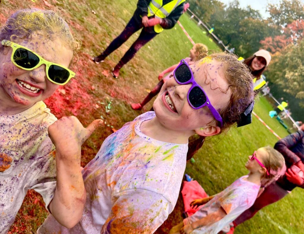 Two young girls, covered in colorful powder and wearing bright sunglasses, smile and one gives a thumbs-up in an outdoor field. Other children and adults are visible in the background.