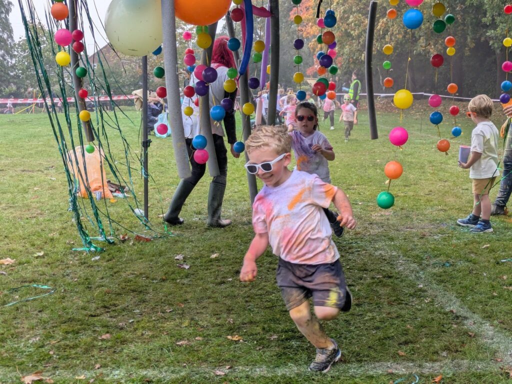 Children, one running in the foreground, participate in an outdoor event on a grassy field decorated with colorful hanging balloons and streamers.
