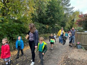 A group of adults and children in school uniforms walk along a tree-lined path, with one child in the foreground holding an art project or container.