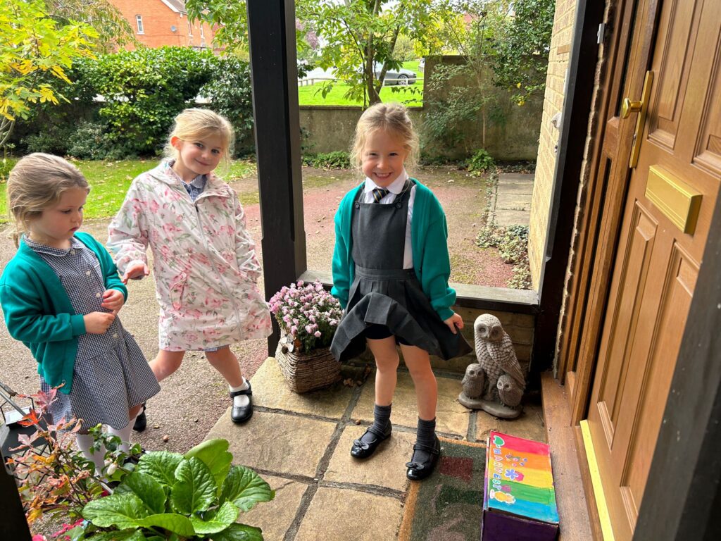 Three young girls in school uniforms stand smiling on a porch outside a front door, one of them posing with a big smile.