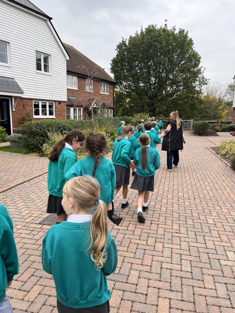 A group of children in teal school tops and grey skirts or shorts walk in a line down a brick-paved path in a residential area, following a woman in a black jacket.