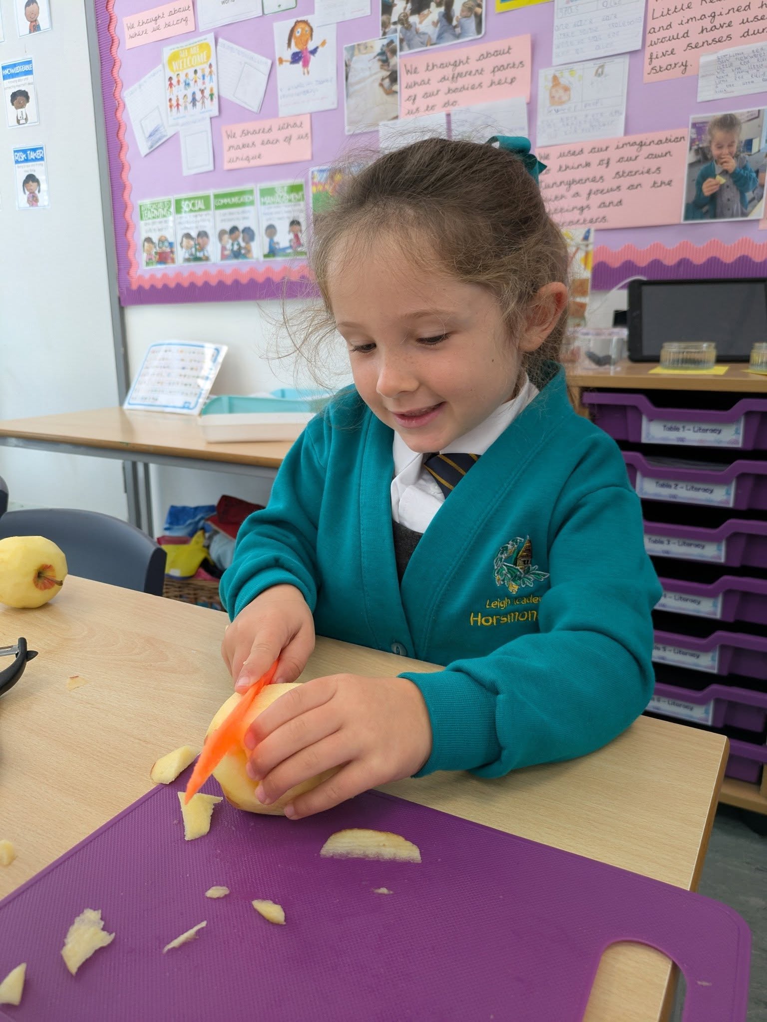 A young girl in school uniform smiling while using a knife to peel an apple on a purple cutting board.