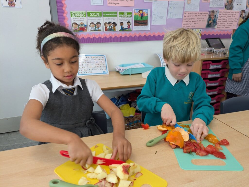 Two children in school uniform cutting up apples and orange peel on cutting boards on a table.