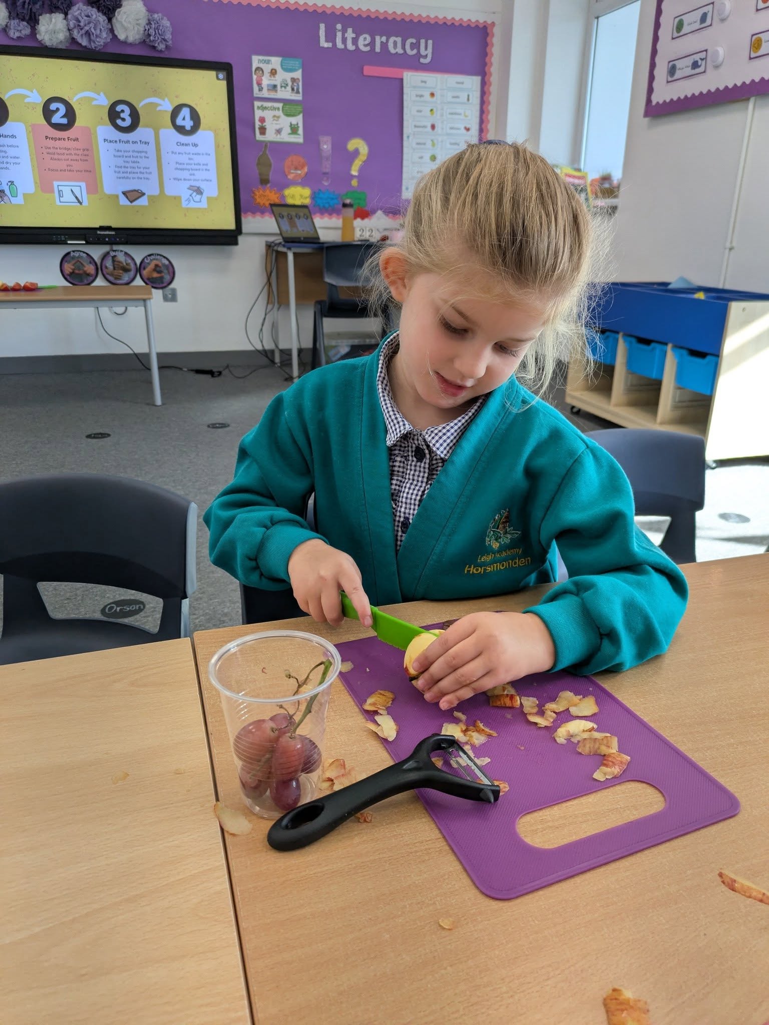 A girl in school uniform peeling an apple with a knife on a purple cutting board, with a cup of grapes nearby.