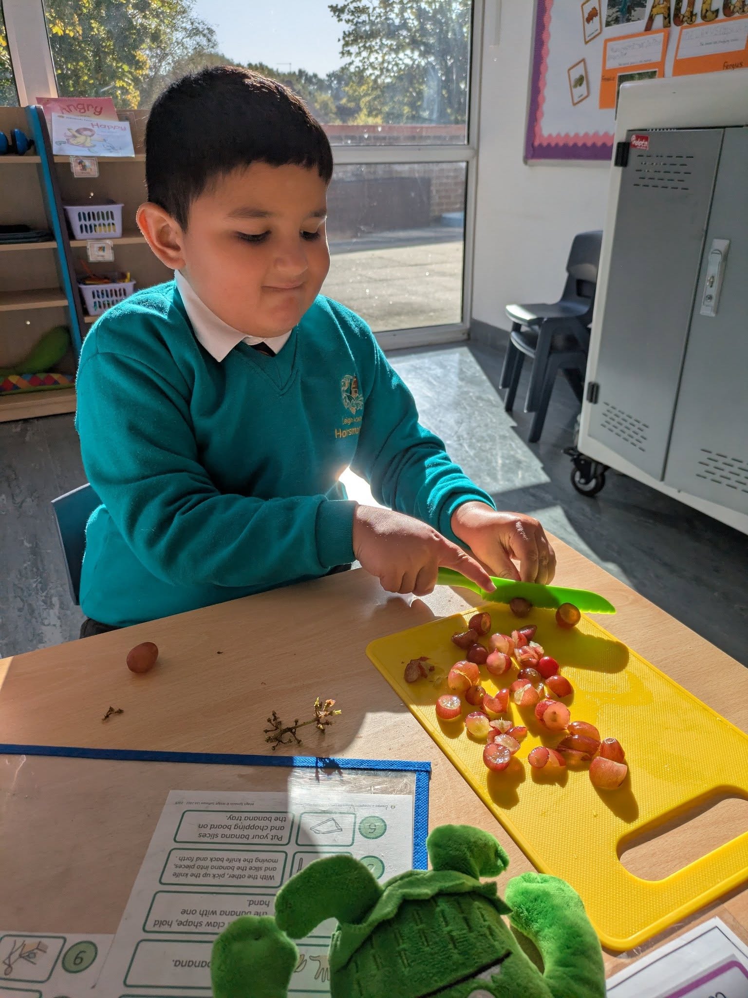 A boy in school uniform carefully cutting grapes on a yellow cutting board at a desk near a window.