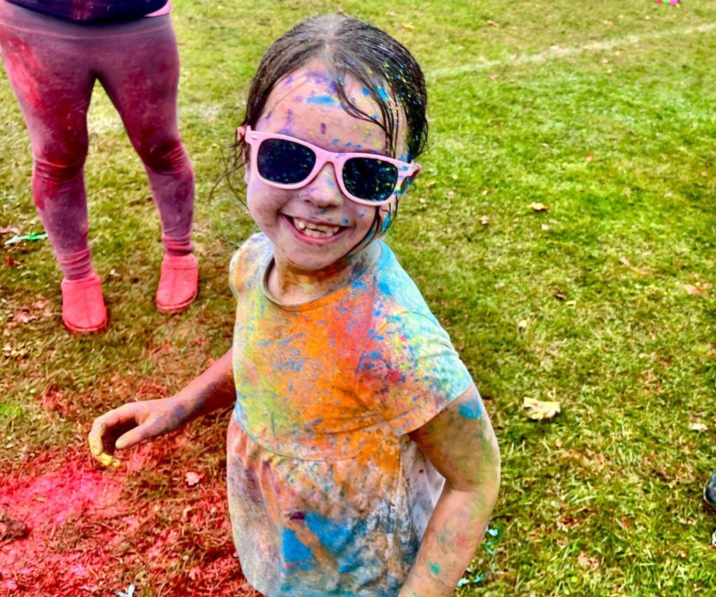 A young girl with colorful powder on her face and clothes smiles while wearing pink sunglasses, standing on a grassy field. A person's legs in pink leggings is partially visible behind her.