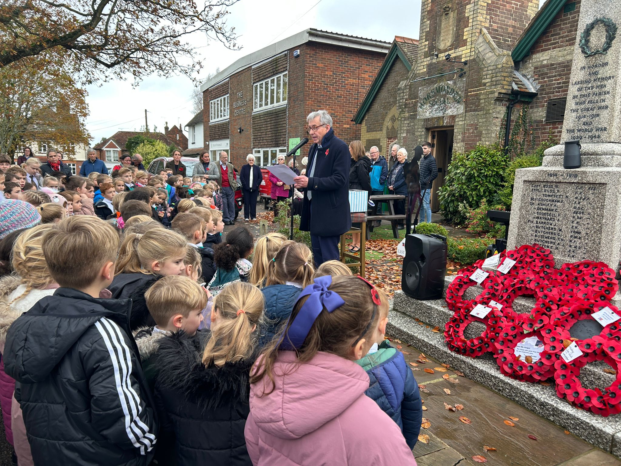 A man speaks into a microphone at a Remembrance Day service near a war memorial covered in poppy wreaths, addressing a large crowd of schoolchildren and adults.