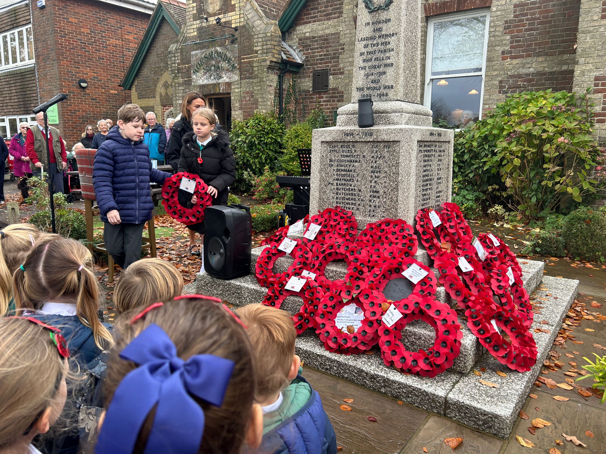 Two schoolchildren stand next to a war memorial adorned with poppy wreaths while a large group of children watches the ceremony.