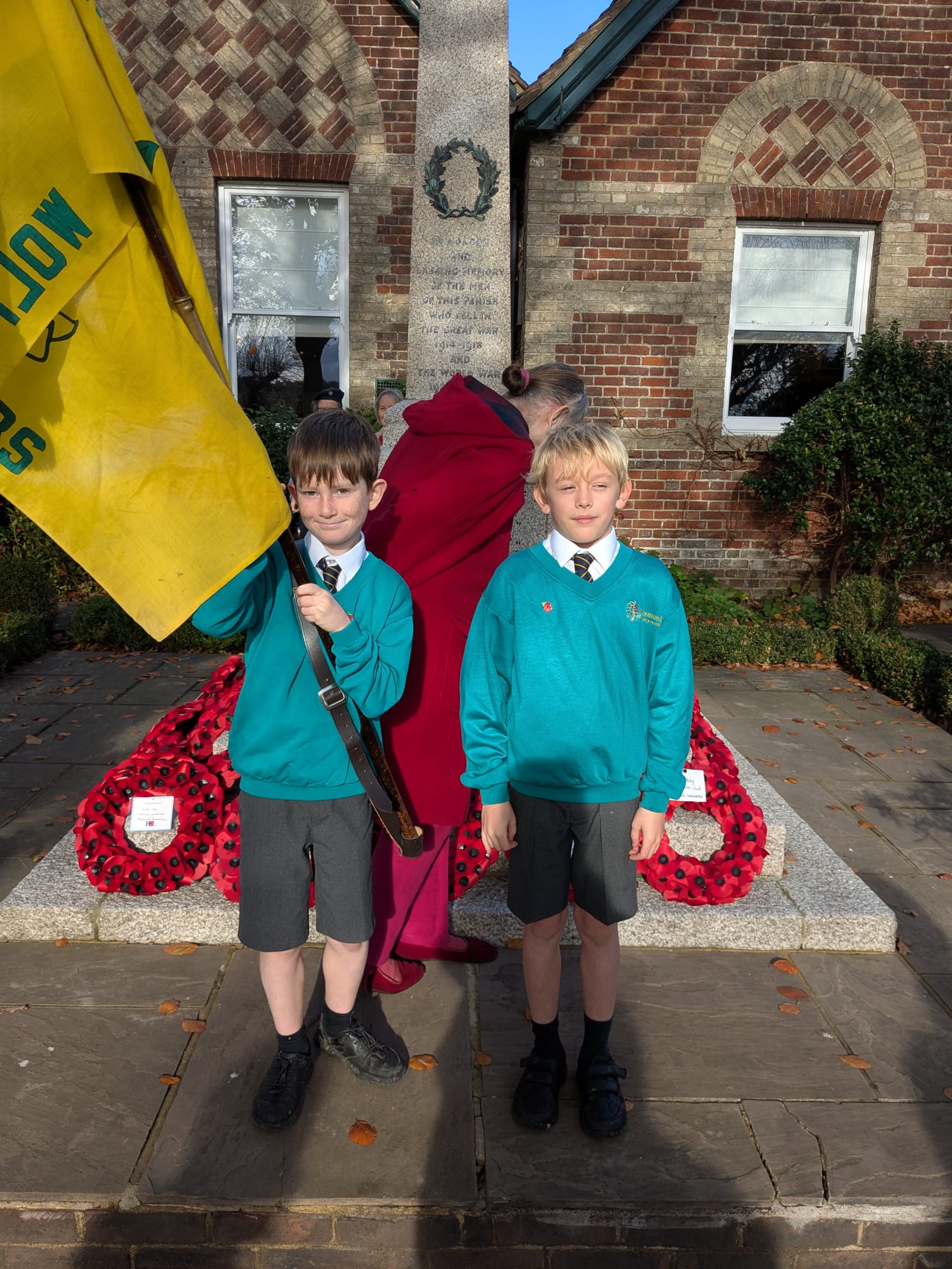 Two boys in school uniforms stand in front of a war memorial covered in poppy wreaths, one holding a yellow flag.