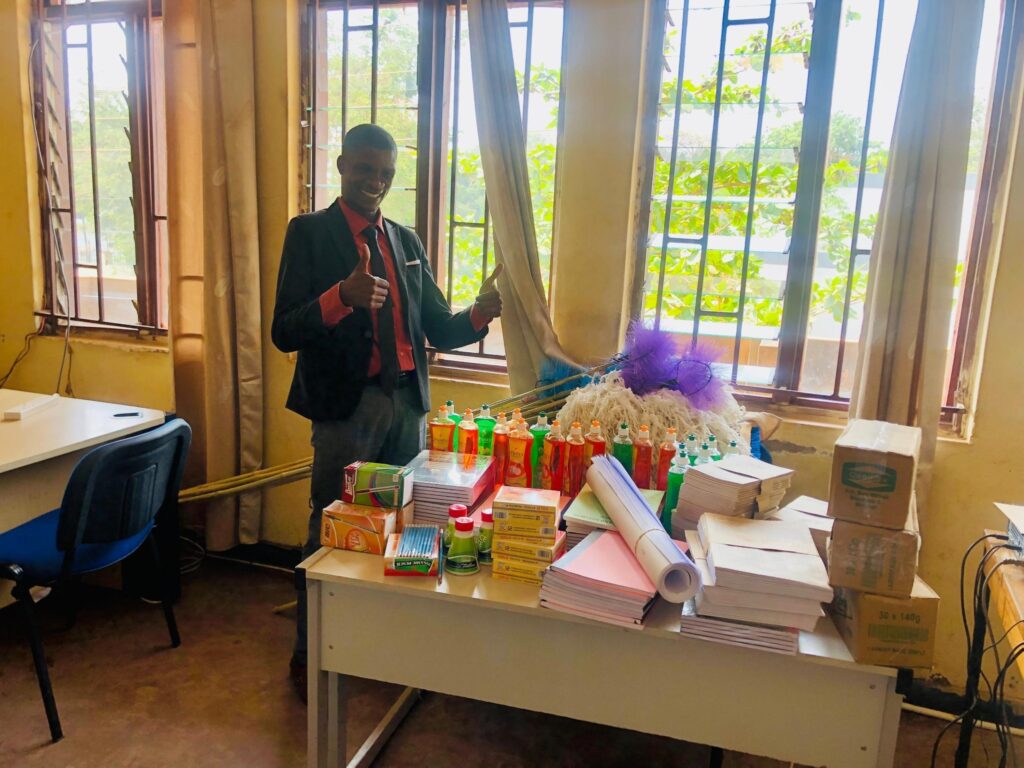 A man in a suit stands next to a desk piled high with school supplies, cleaning supplies, and other materials, giving a thumbs-up gesture.