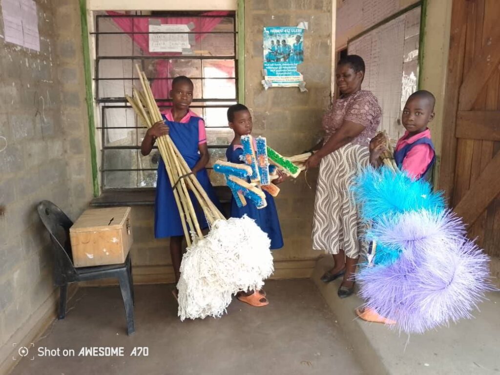 A woman and three girls in school uniforms stand next to a doorway holding various cleaning tools, including brooms and scrubbing brushes.