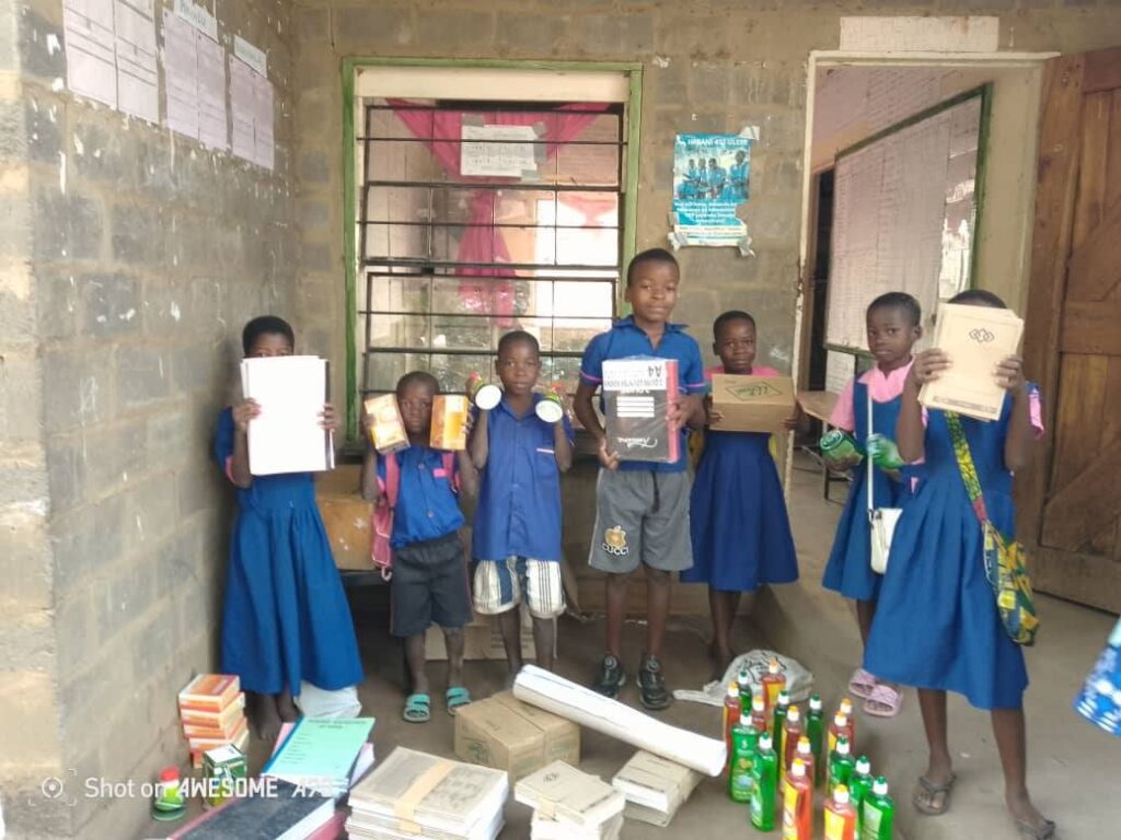 Six children, dressed in school uniforms and casual clothes, stand in a simple room holding various school supplies and cleaning products. There are stacks of books and bottles on the floor in front of them.