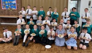 A large group of children in school uniforms are posing indoors, holding small loaves of bread or baked goods on plates.