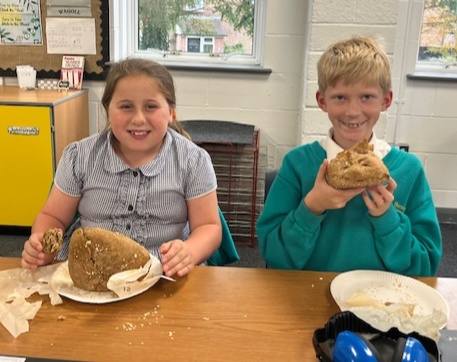 Two smiling children, wearing school uniforms, sit at a table holding and looking at small loaves of bread or baked goods they have made.