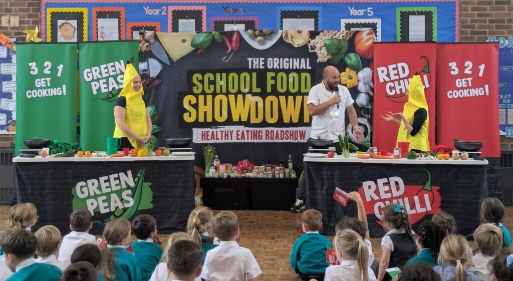 An indoor "School Food Showdown" event with two cooking stations—labeled "Green Peas" and "Red Chilli"—is being presented by a chef and two people in banana costumes to an audience of schoolchildren.