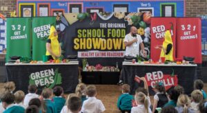 An indoor "School Food Showdown" event with two cooking stations—labeled "Green Peas" and "Red Chilli"—is being presented by a chef and two people in banana costumes to an audience of schoolchildren.