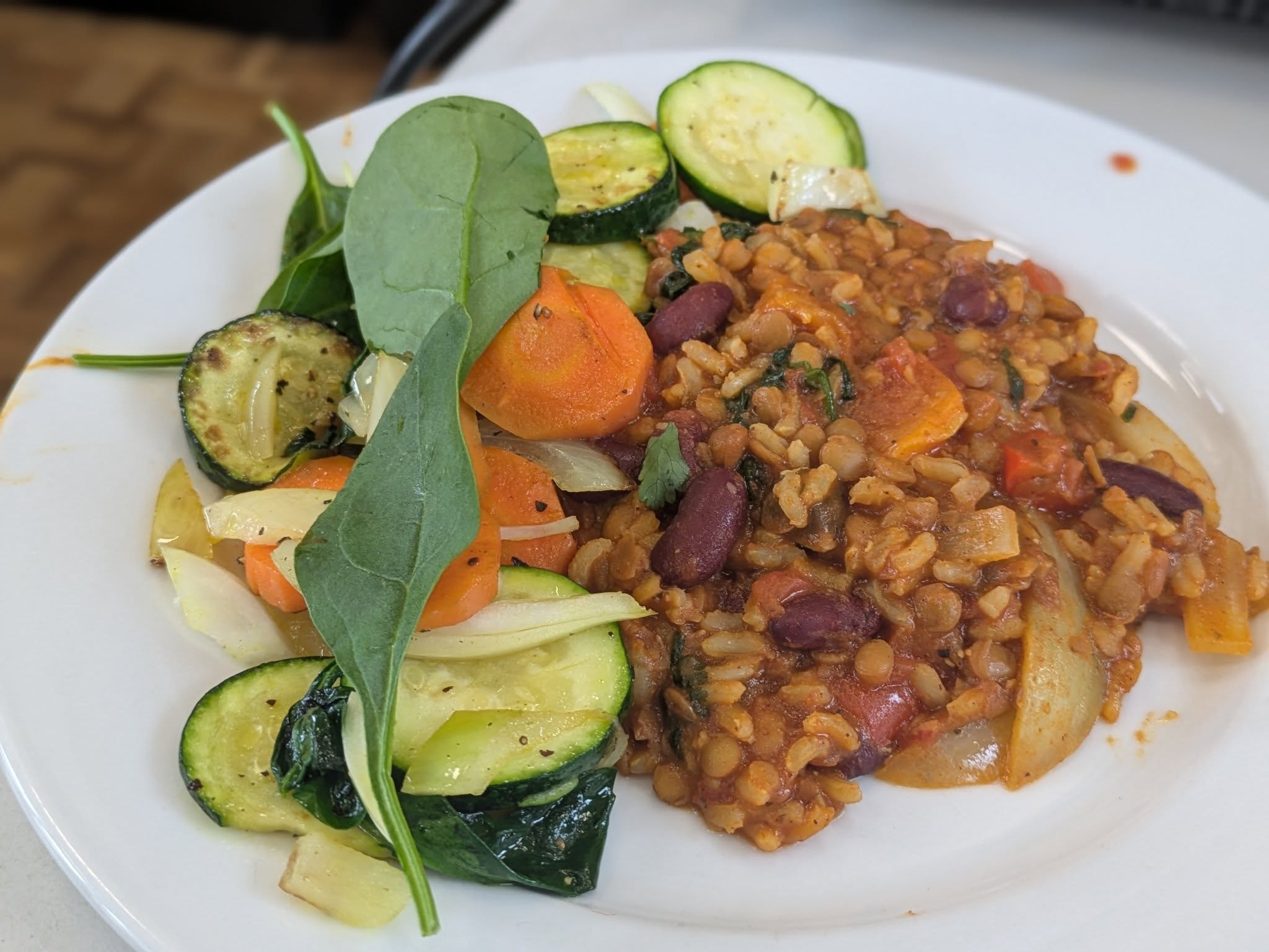 A white plate holds a serving of lentil and bean stew alongside sautéed vegetables, including zucchini, carrots, and onions, garnished with spinach leaves.